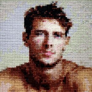 Dreamboat — original 4×4 inch hand-stitched cross-stitch portrait of a young dark-haired man with a direct gaze, stitched in warm brown, cream, and blush tones against a soft sage and lavender background